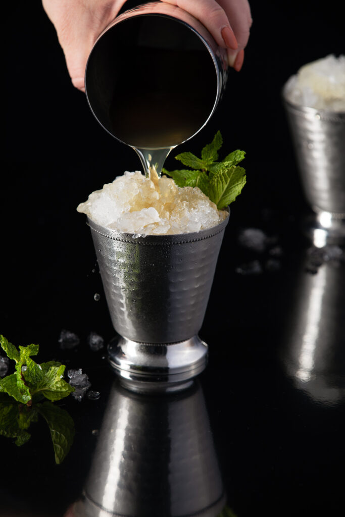 A white hand pours a mint julep into a silver julep cup.