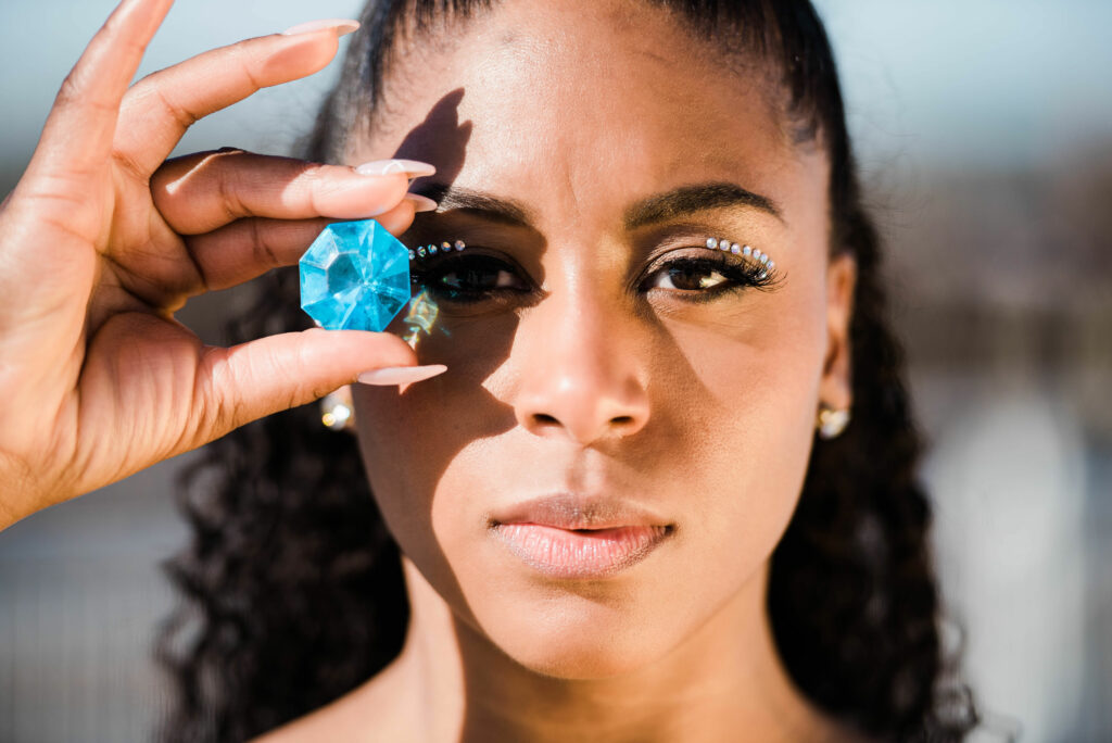 A black woman holds a blue gem up to her eye and looks directly at the camera.