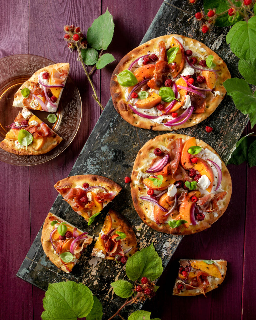 An aerial view of three Peach Wineberry Flatbreads, with one cut into six slices. They are bright in color and sit on a black wooden slab. Peach Wineberry Flatbread Recipe