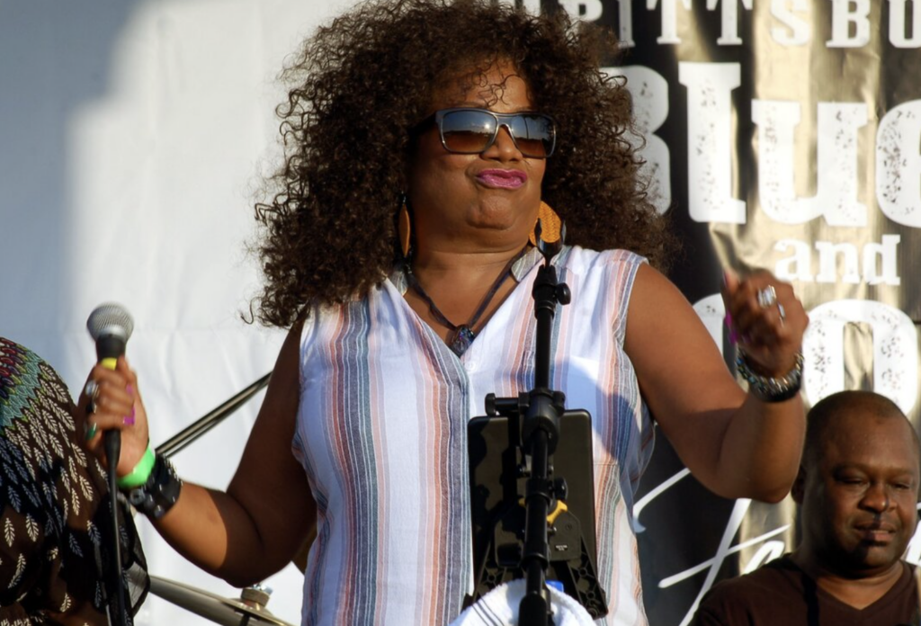 A black woman stands on a stage with a microphone in her right hand.