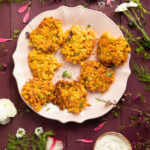 An aerial view of seven Sweet Potato and Corn Fritters on an a white plate, surrounded by herbs. Corn Fritters recipe. Vegetable fritter