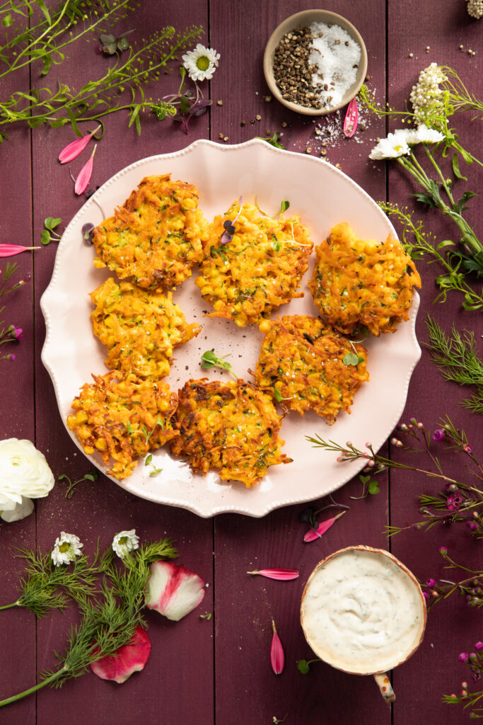 An aerial view of seven Sweet Potato and Corn Fritters on an a white plate, surrounded by herbs. Corn Fritters recipe. Vegetable fritter
