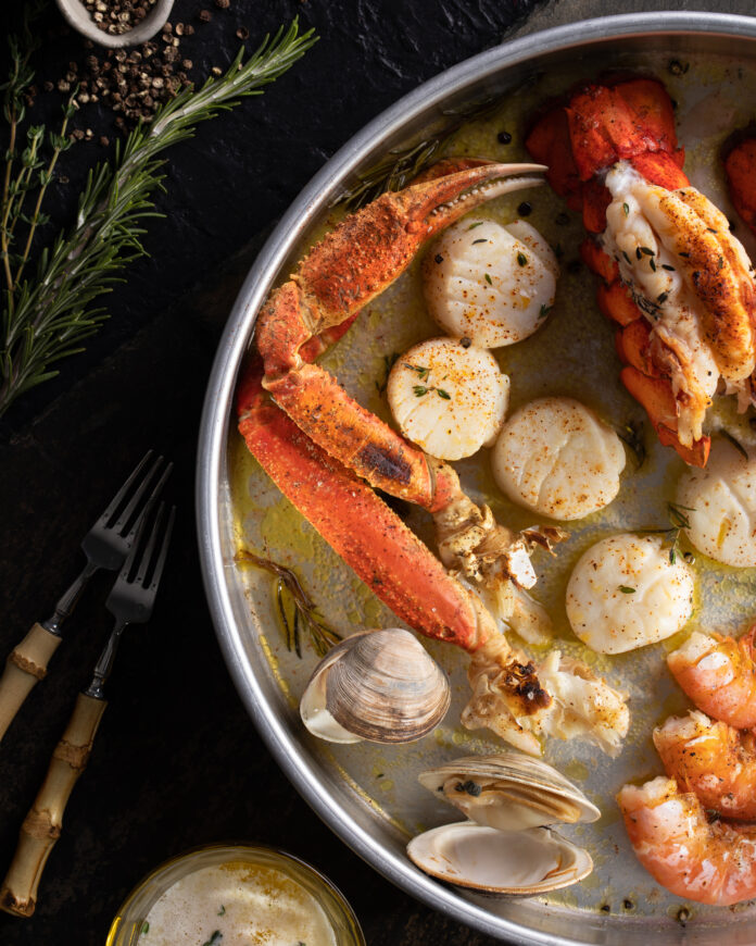 Seafood Broil for 2 a black table with a round metal pan with clams, scallops, crab legs, lobster, and shrimp