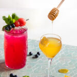 a look in on a light green tile surface and white background of a dark pink cocktail in a tall glass with mint, blueberry and watermelon garnish, and a honey yellow cocktail tin a stemmed glass garnished with honeycomb and a drip of honey from a honey stir stick held by a woman. Beyoncé cocktail