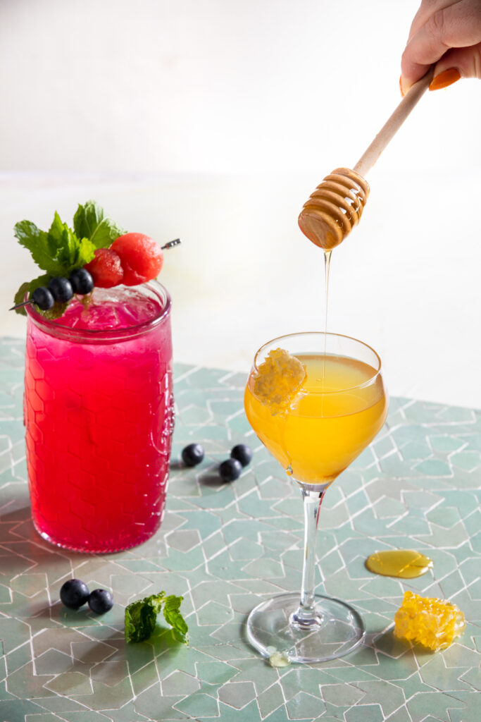 a look in on a light green tile surface and white background of a dark pink cocktail in a tall glass with mint, blueberry and watermelon garnish, and a honey yellow cocktail tin a stemmed glass garnished with honeycomb and a drip of honey from a honey stir stick held by a woman. Beyoncé cocktail