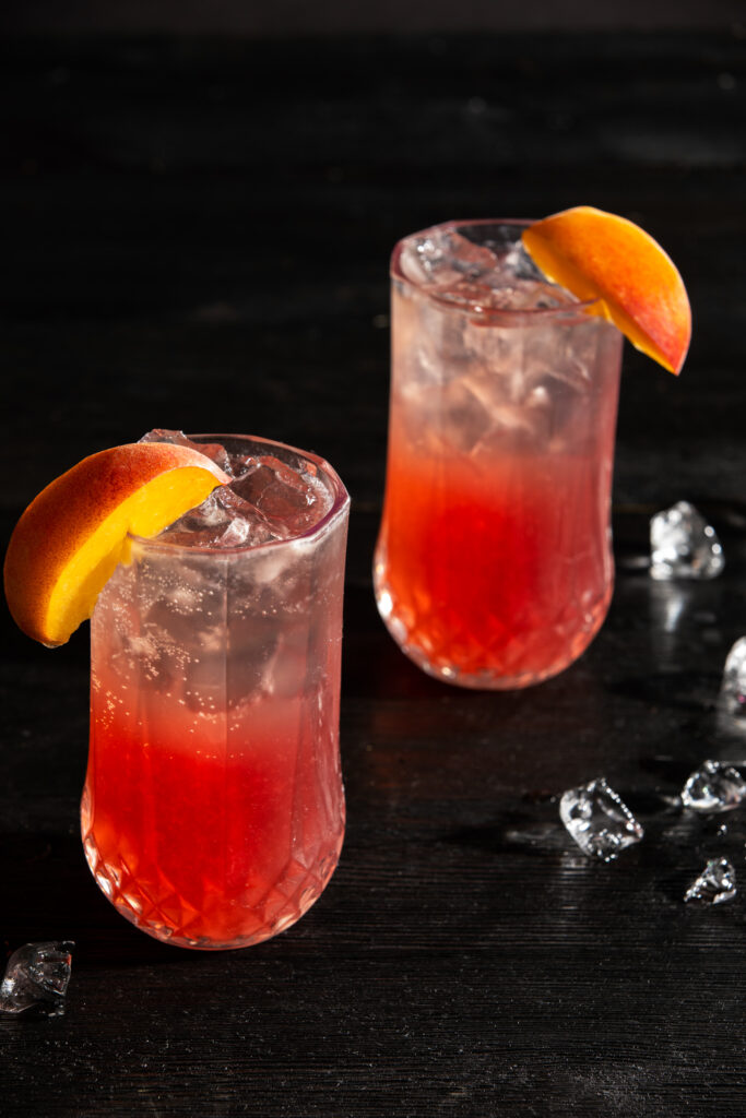 a look in at 2 identical dark peachy pink cocktail on a black background with ice cube and peach wedges as garnish