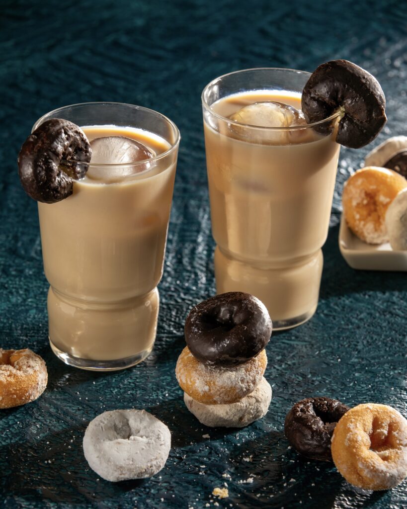 two cocktails that have a light brown color with round cocktail ice cubes and mini cake, powdered and chocolate coated donuts as garnish