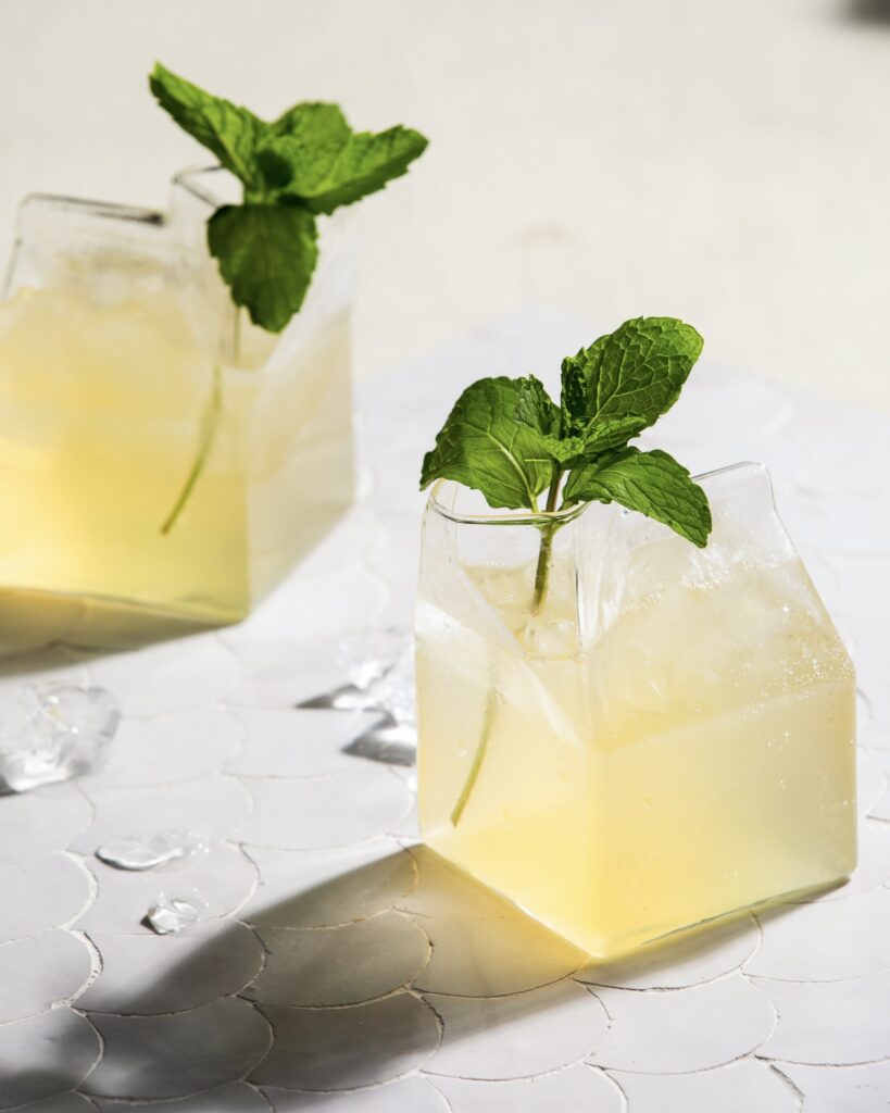 two yellow cocktails in small glass cartons with mint sprig garnish on a white tile surface