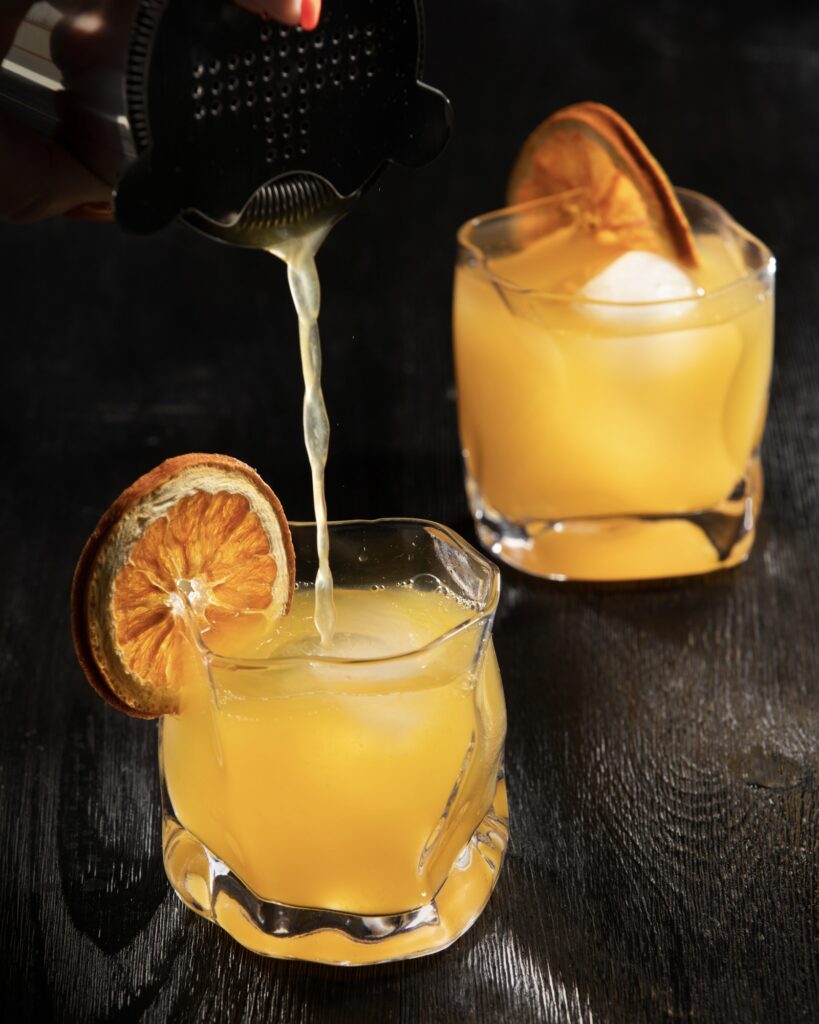 2 orange cocktail in rocks glasses on a black surface with dehydrated orange slices as garnish. The front cocktail is being poured by a woman's hand
