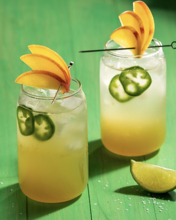 2 glasses with peach colored cocktails garnished with peach slice fans and jalapeño slices sitting on a vibrant green wooden surface