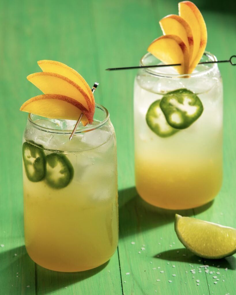 2 glasses with peach colored cocktails garnished with peach slice fans and jalapeño slices sitting on a vibrant green wooden surface