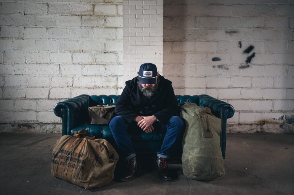 A man with a hat and a beard sits on a leather couch and looks down. He's surrounded by duffle bags.