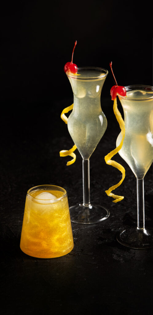 a look in photo with a black background and 3 cocktail glasses. 2 are the shape of a woman's body with a very pale yellow cocktail garnished with a long lemon twist and maraschino cherry, and a single glittery gold cocktail in a shorter glass