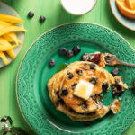 A stack of blueberry pancakes on a green plate. A forkful is taken out of the stack, with the fork sitting to the top right of the pancakes. Blueberry Pancakes Recipe