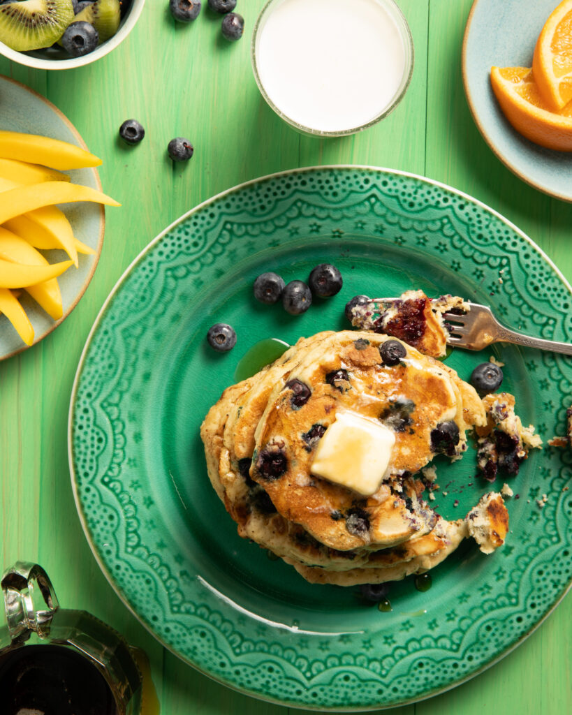 A stack of blueberry pancakes on a green plate. A forkful is taken out of the stack, with the fork sitting to the top right of the pancakes. Blueberry Pancakes Recipe