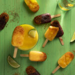 Various dark red and orange colored fruit popsicles sit on green wood. Paleta Popsicles Recipe