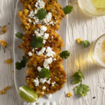 4 corn fritters topped with cotija cheese and cilantro, and garnished with a lime wedge on a narrow white oval platter on a white surface with 2 glasses of beer on the right side