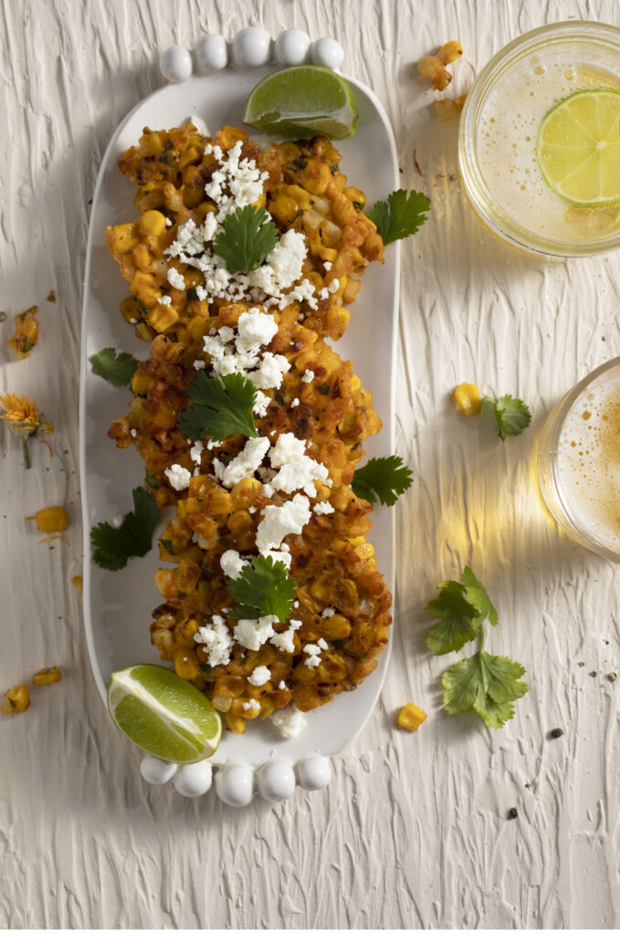 4 corn fritters topped with cotija cheese and cilantro, and garnished with a lime wedge on a narrow white oval platter on a white surface with 2 glasses of beer on the right side