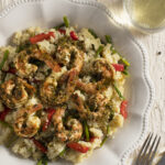 A large scalloped edge white dish with shrimp over couscous, and a glass of wine on a textured white surface