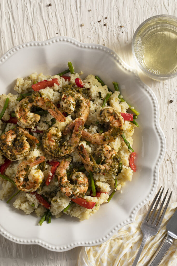 A large scalloped edge white dish with shrimp over couscous, and a glass of wine on a textured white surface