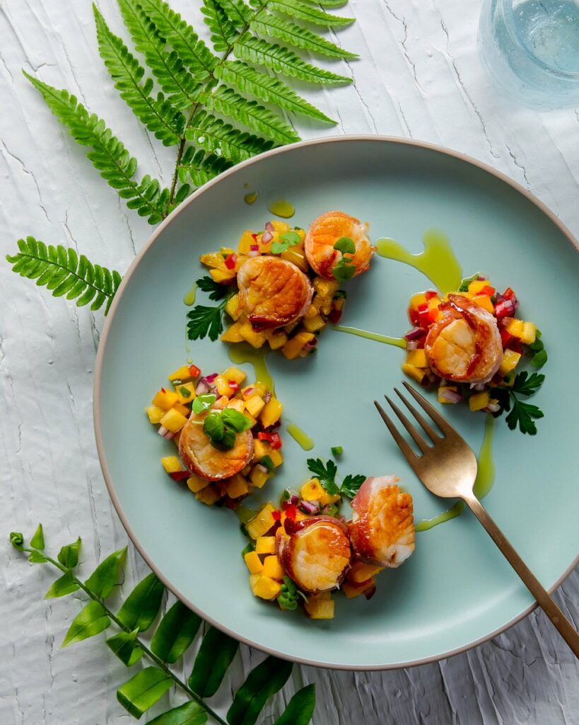 Four bacon wrapped scallops with fruity mango pineapple salsa sits on a blue plate.
