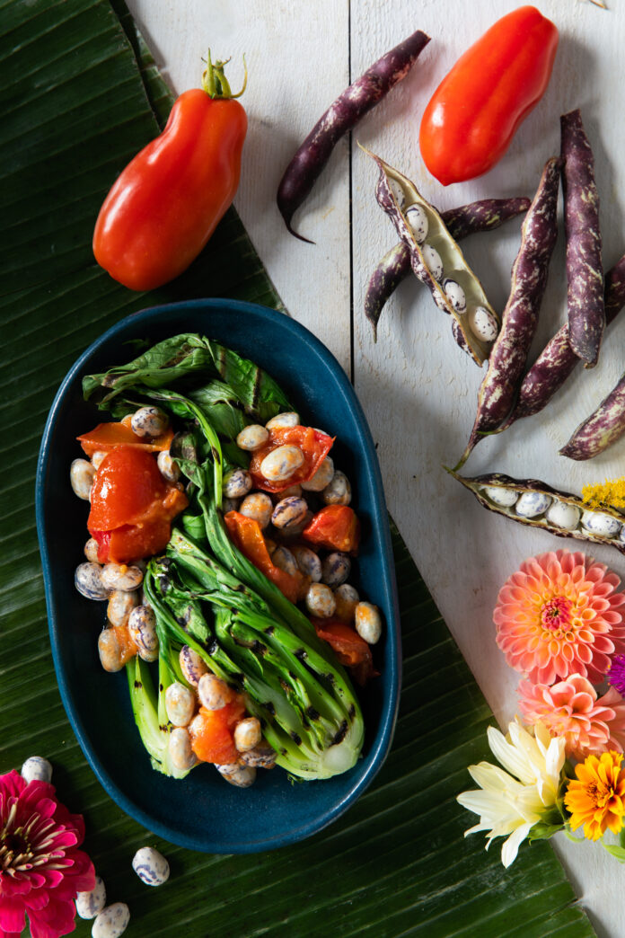 A flat-lay image of a brightly colored dish of beans and greens with colorful flowers and vegetables.