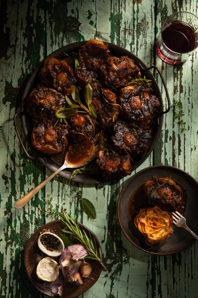 Flat-lay image of Green Chile Braised Oxtail in a cast-iron pan set on a weathered table. Braised Oxtail Recipe