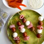 An aerial shot of four Caprese Kebabs sitting on a green plate. A bowl of carrots and dip sit nearby. Caprese Kebabs Recipe