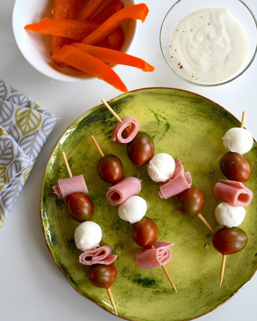 An aerial shot of four Caprese Kebabs sitting on a green plate. A bowl of carrots and dip sit nearby. Caprese Kebabs Recipe