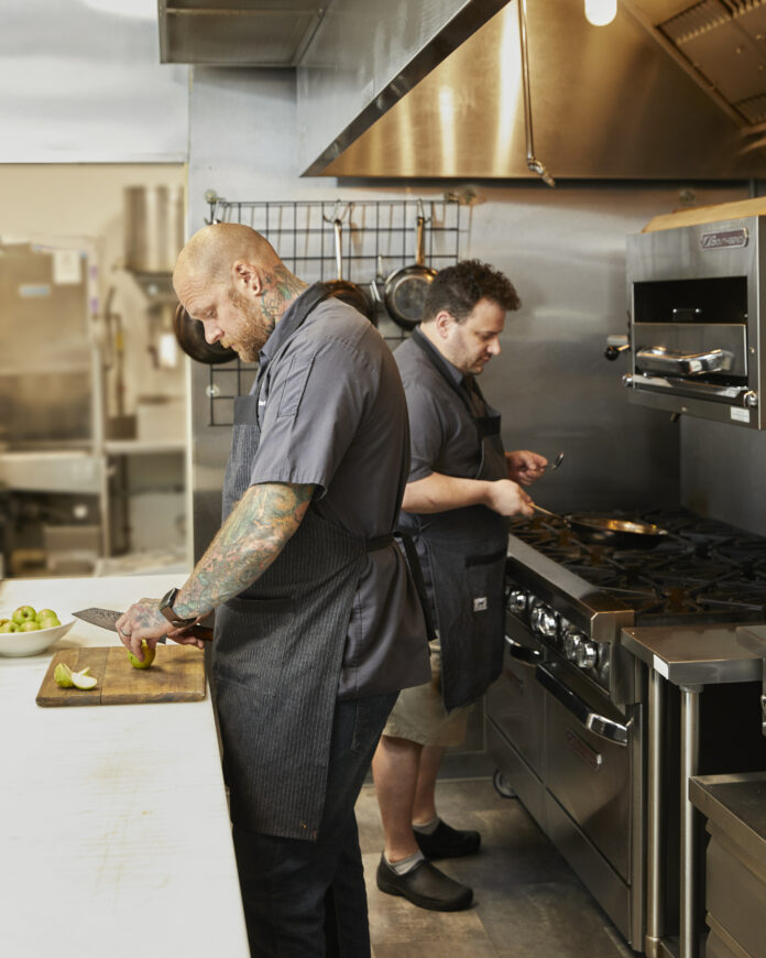 Chef Cory Hughes, wearing a black chefs outfit, its bent over a counter in Fig and Ash's kitchen cutting veggies.
