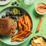 An aerial view of a Turkey and Black Bean Burger, Sweet Potato Fries, blueberrys, and kiwi slices sitting on a light green plate against a green background.