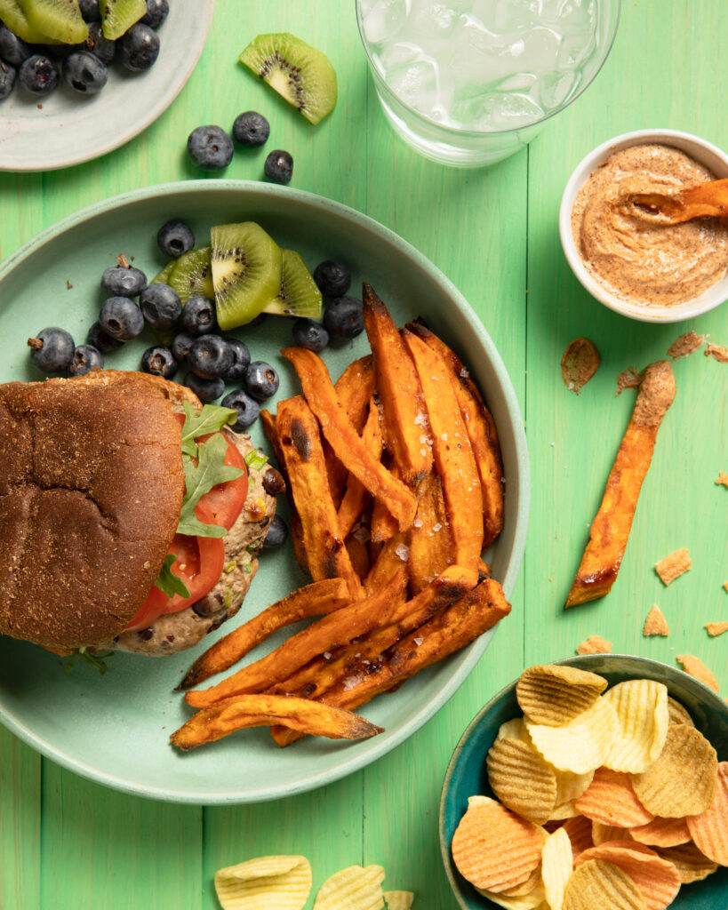 An aerial view of a Turkey and Black Bean Burger, Sweet Potato Fries, blueberrys, and kiwi slices sitting on a light green plate against a green background.