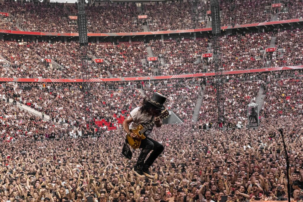 A man in a tall hat jump with a guitar in front of a stadium crowd.