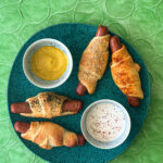 Four hot dog roll-ups sit on a green plate agasint a green backgrouns