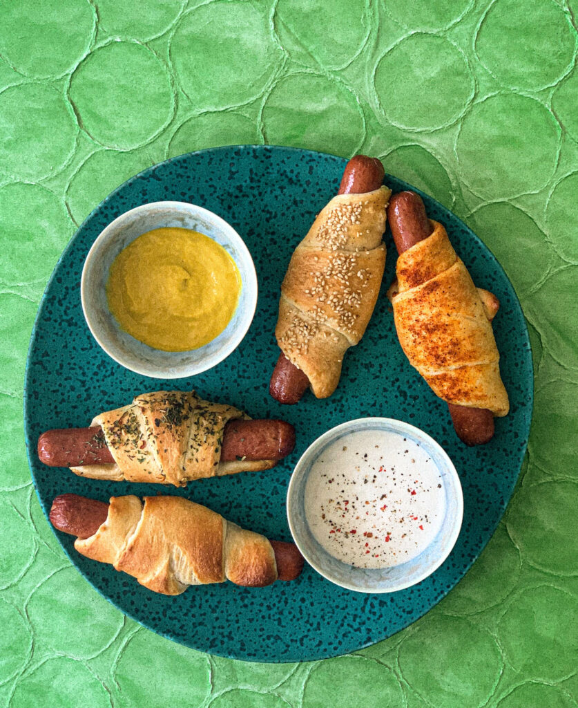 Four hot dog roll-ups sit on a green plate agasint a green backgrouns