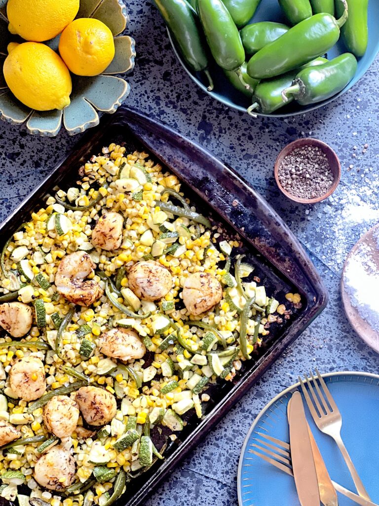 A sheet pan filled with Summer Scallops and Corn. To the side sits bowls of peppers, lemon, and salt. Scallops recipe