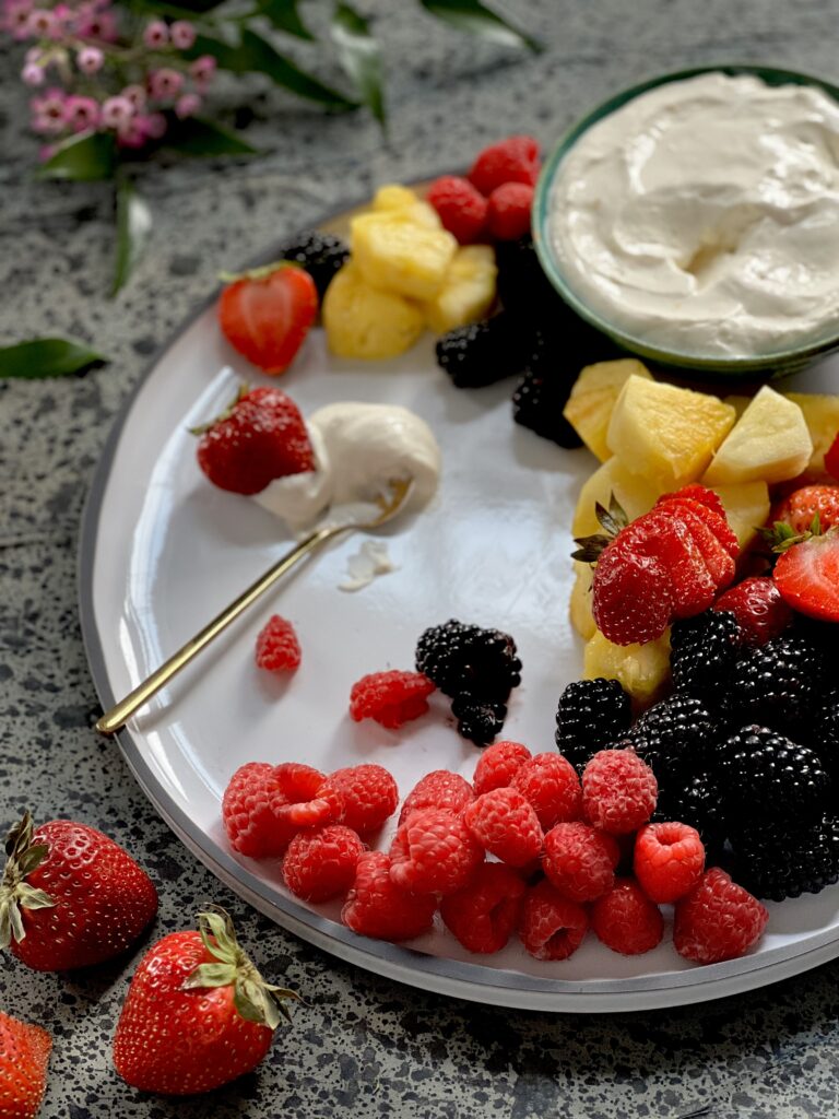 Berries and pineapple on a white plate with a bowl of creamy white fruit dip, and a little gold spoon with some of the dip on a plate