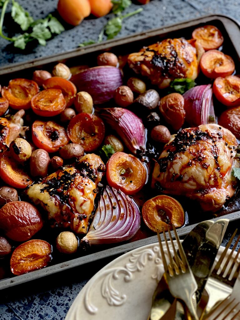 A sheet pan with chicken thighs, halved apricots, purple onions and baby potatoes with a stack of creamy yellow plates and gold forks. Asian Stone Fruit Chicken Bake Recipe