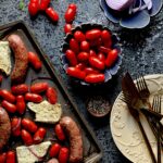 A sheet pan dinner with sausage, red grape tomatoes, and feta cheese with a bowl of tomatoes and a plate with red onion