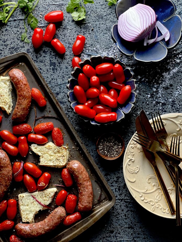A sheet pan dinner with sausage, red grape tomatoes, and feta cheese with a bowl of tomatoes and a plate with red onion