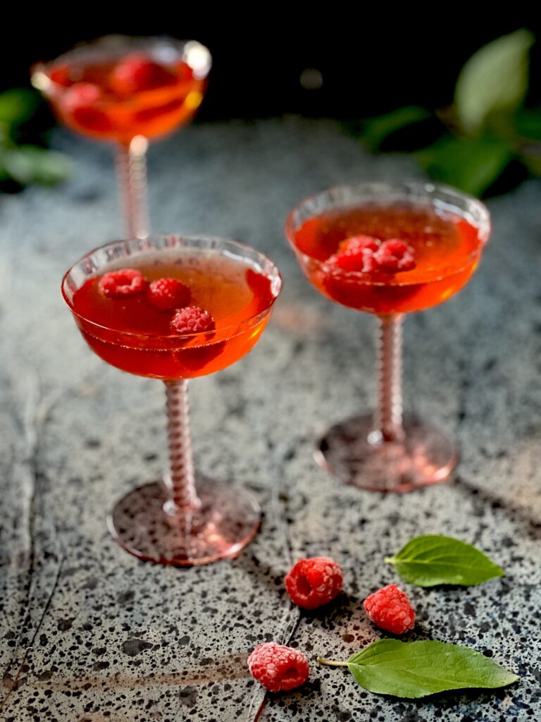 Three dark pink cocktails in pink coupe glasses with floating raspberries