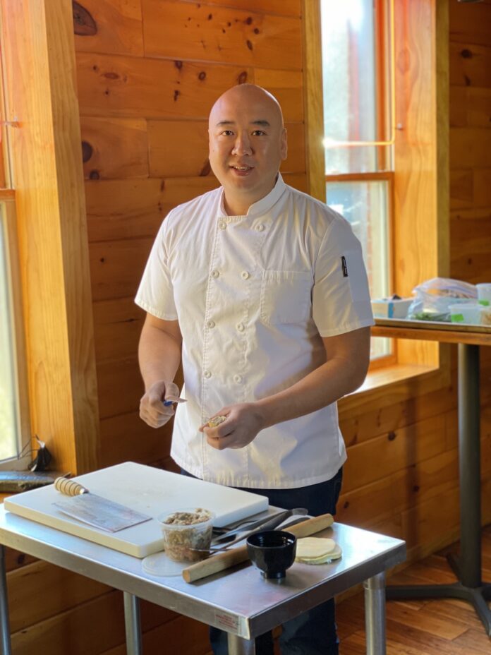Chef Roger Li Chef Roger Li, in a white chefs jacket, stands facing the camera over a small prep table.