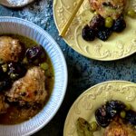 An arial view of a round white baking dish with chicken thighs, olives, prunes, and capers, and two yellow dishes with single servings of the same with gold forks
