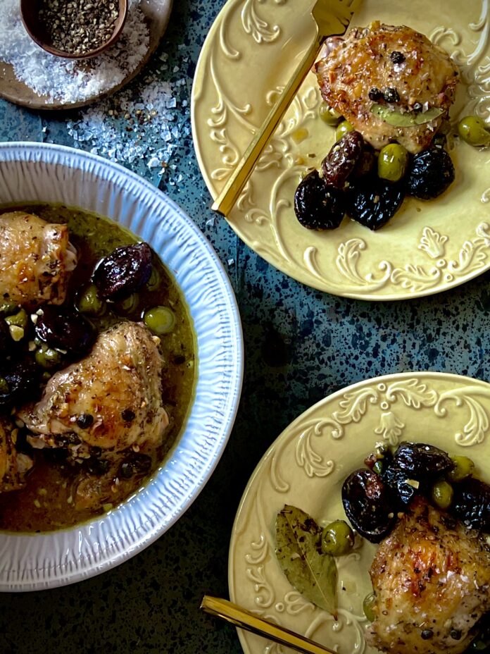 Chicken Marbella An arial view of a round white baking dish with chicken thighs, olives, prunes, and capers, and two yellow dishes with single servings of the same with gold forks