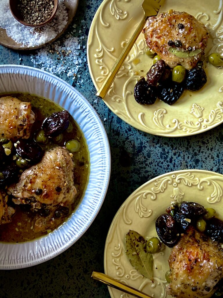 An arial view of a round white baking dish with chicken thighs, olives, prunes, and capers, and two yellow dishes with single servings of the same with gold forks