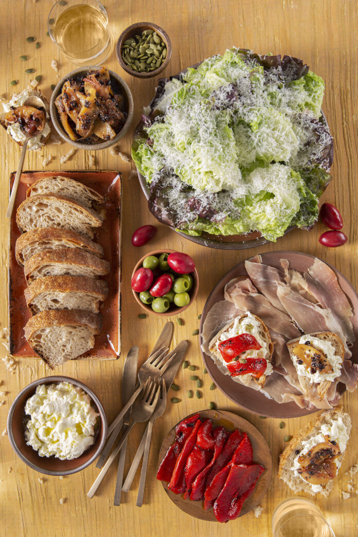 An aerial view of a fall dinner menu spread featuring bread, meats, tomatoes, a fresh salad sprinkled with cheese, and more.