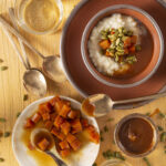 Rice Pudding with Candied Pumpkin and Caramel sits in an orange and brown bowl. Bowls of candied pumpkin, and caramel sit below the pudding. Rice Pudding recipe