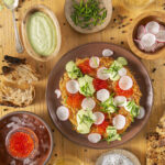 A colorful Potato Rösti with smoked trout roe, crème fraîche, and dill is artfully arranged on a brown plate. Surrounding the plate are small bowls of the garnishes: Crème fraîche, Chopped dill. Chopped chives, and Trout roe. Potato Rösti recipe