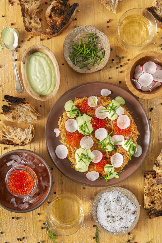 A colorful Potato Rösti with smoked trout roe, crème fraîche, and dill is artfully arranged on a brown plate. Surrounding the plate are small bowls of the garnishes: Crème fraîche, Chopped dill. Chopped chives, and Trout roe. Potato Rösti recipe