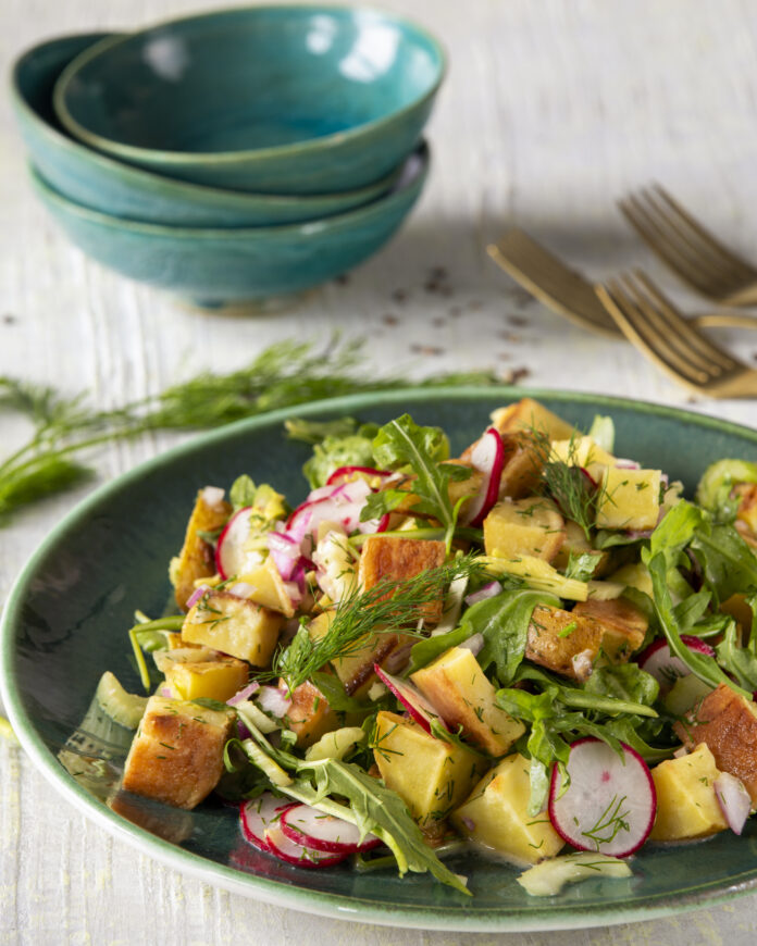 Three individual blue bowls above a green serving bowl filled with a vibrant potato salad of yellow, green, red, and purple ingredient